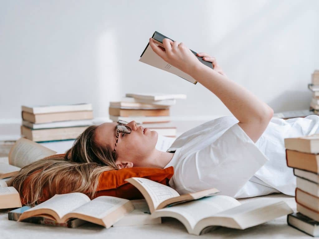 Temas para tesis: estudiante investigando y leyendo libros para inspirarse en su proyecto académico Estudiante universitaria acostada leyendo un libro, rodeada de libros abiertos y apilados, buscando inspiración para elegir temas de tesis.